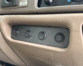 May include: A black panel with four illuminated blue rocker switches, mounted on a tan dashboard. The switches are evenly spaced and have a small blue light in the center. The dashboard also features other vehicle controls.