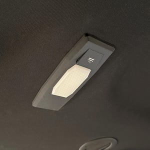 Puede incluir: Una lámpara de techo negra con una luz rectangular blanca y texturizada y un pequeño interruptor con un icono de bombilla. La lámpara está montada sobre una superficie gris oscura y texturizada.