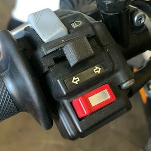 May include: Close-up of a motorcycle handlebar with black controls and a red button. The controls include a gray switch, a headlight switch, and turn signal indicators.