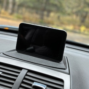 May include: A black tablet device is mounted on a dashboard in a car. The tablet is positioned vertically and is facing the driver. The tablet is likely being used for navigation or entertainment.