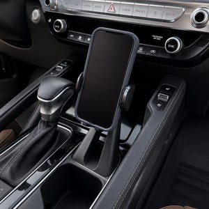 May include: A black smartphone mounted in a vertical car phone holder. The holder is secured within the center console cup holder area of a vehicle, positioned between the gear shift lever and the dashboard controls with silver accents.