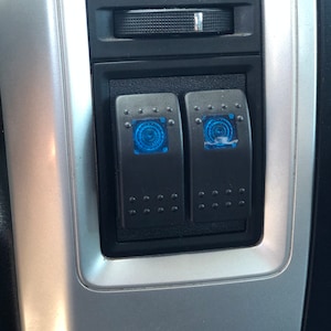 May include: Two black toggle switches with blue LED lights. The switches are mounted in a silver panel.