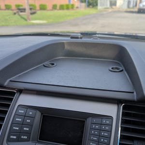 Puede incluir: Primer plano del salpicadero de un coche. El salpicadero es negro con una superficie texturizada e incluye una radio, un reproductor de CD y otros controles. El salpicadero tiene dos aberturas circulares y un área de almacenamiento rectangular.