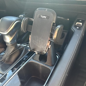 May include: Black TOPK phone holder for a car, mounted in the center console. The holder has adjustable arms and a textured surface. The car interior is black and silver.