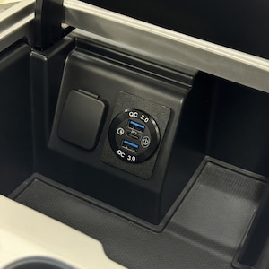 Könnte beinhalten: Schwarze Autokonsole mit einer Steckdose und einer USB-Ladestation. Die USB-Station hat zwei Anschlüsse mit der Aufschrift "QC 3.0" und einen Power-Knopf. Die Konsole ist geöffnet und zeigt ein Staufach.