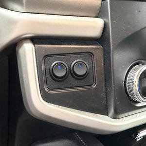 Puede incluir: Primer plano del salpicadero de un vehículo con dos interruptores negros y circulares con luces indicadoras azules. Los interruptores están colocados en un panel negro rectangular, rodeado de un embellecedor plateado y negro.