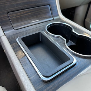 May include: A close-up of a car's center console with a black plastic storage compartment, two cup holders, and a wood-grain finish.