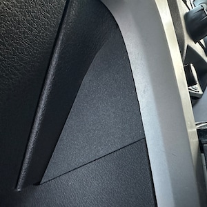 May include: A close-up shot of a car's interior, showing textured black plastic panels and a light grey trim. The image focuses on the contrasting textures and shapes within the vehicle's design.