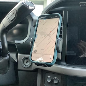Può includere: Un supporto per telefono nero e grigio attaccato alla bocchetta di ventilazione di un'auto. Uno smartphone con un'app di mappe aperta è nel supporto. Lo schermo del telefono mostra una mappa di un quartiere con il testo "Waesila" visibile.