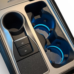 Puede incluir: Consola central de coche con portavasos con iluminación azul y botón de freno de estacionamiento. La consola tiene una combinación de colores plateado y negro, con una superficie negra texturizada cerca de los botones.