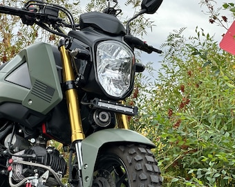 May include: Close-up of a matte olive green motorcycle with gold suspension forks. The front features a round headlight, a small LED light bar, and a black and gray American flag graphic. The tire has a rugged tread.