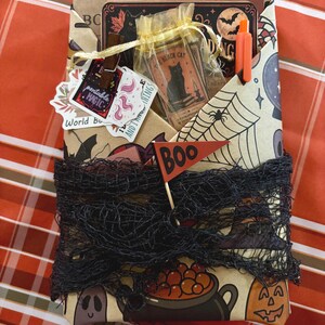 May include: A rectangular gift package wrapped in tan paper featuring illustrations of ghosts, pumpkins, and cauldrons. It is wrapped in black netting and adorned with a red pennant flag labeled "Boo," a gold organza bag, stickers, and an orange pen.