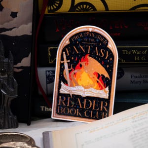 Puede incluir: Una pegatina con un diseño negro y dorado que presenta un dragón que emerge de un libro con una espada. El texto dice "Proud Member of the Fantasy Reader Book Club".