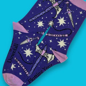 May include: A pair of purple socks with a pattern of weapons and stars. The socks have light purple cuffs and toes. The text "Downfall" and "Plotting Your" are printed on the socks.