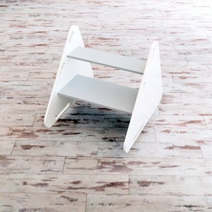 May include: A white wooden step stool with two gray steps. The stool has a modern design with triangular side supports and is suitable for reaching higher shelves or surfaces. The stool is on a light wood floor.