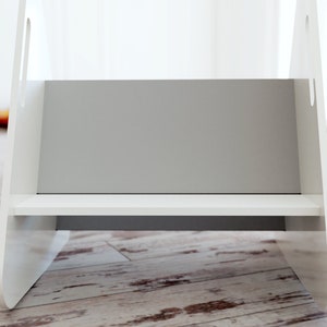 May include: A white wooden step stool with a grey interior and a single shelf. The stool has cut-out handles on the sides and is designed for children or to reach higher places. The step stool is on a wooden floor.