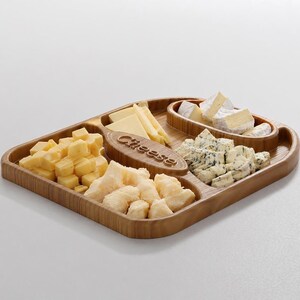 Pode incluir: Uma t&aacute;bua de queijos de madeira com v&aacute;rios queijos, incluindo cubos, fatias e esfarelados. A t&aacute;bua tem uma pega embutida e compartimentos. A palavra "Cheese" est&aacute; gravada no centro da t&aacute;bua.