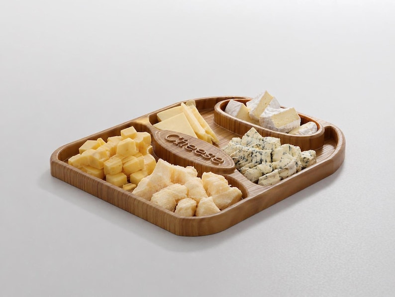 Pode incluir: Uma t&aacute;bua de queijos de madeira com v&aacute;rios queijos, incluindo cubos, fatias e esfarelados. A t&aacute;bua tem uma pega embutida e compartimentos. A palavra "Cheese" est&aacute; gravada no centro da t&aacute;bua.