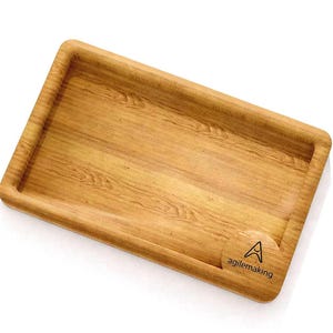 K&ouml;nnte beinhalten: Ein rechteckiges Holztablett mit erh&ouml;htem Rand und nat&uuml;rlicher Holzmaserung. Das Tablett hat abgerundete Ecken und eine kleine kreisf&ouml;rmige Vertiefung mit dem Text "agilemaking" und einem Logo.