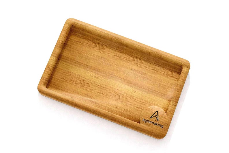 K&ouml;nnte beinhalten: Ein rechteckiges Holztablett mit erh&ouml;htem Rand und nat&uuml;rlicher Holzmaserung. Das Tablett hat abgerundete Ecken und eine kleine kreisf&ouml;rmige Vertiefung mit dem Text "agilemaking" und einem Logo.