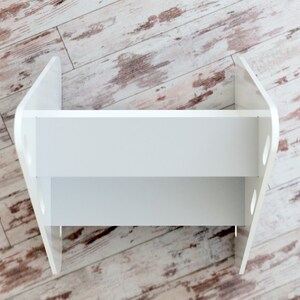 May include: A white, two-tiered shelf unit with a distinctive, curved design. The shelf has a rectangular shape with rounded edges and cut-out details on the sides. The unit is made of a solid, opaque white material, suitable for displaying items.