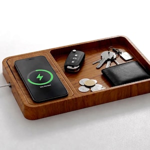 Op de afbeelding: Een houten oplaadstation met een ingebouwde draadloze oplader. Een smartphone laadt links op en toont een groen oplaadsymbool. De lade bevat sleutels, een portemonnee, munten en een autosleutelhanger. Het hout heeft een natuurlijke nerf.