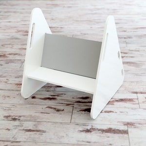 May include: A white wooden step stool with a gray panel in the center. The step stool has a simple design with triangular sides and a rectangular step. It features cut-out handles on the sides.