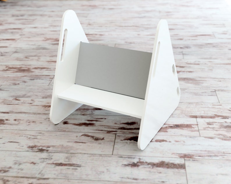 May include: A white wooden step stool with a gray panel in the center. The step stool has a simple design with triangular sides and a rectangular step. It features cut-out handles on the sides.