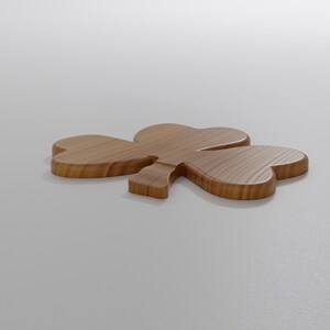 May include: A wooden shamrock, a symbol of St. Patrick's Day, lies on a white surface. The shamrock is made of light brown wood with visible grain, featuring three heart-shaped leaves and a stem. The object is positioned at a slight angle.