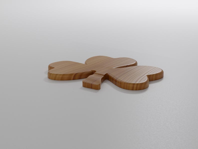 May include: A wooden shamrock, a symbol of St. Patrick's Day, lies on a white surface. The shamrock is made of light brown wood with visible grain, featuring three heart-shaped leaves and a stem. The object is positioned at a slight angle.