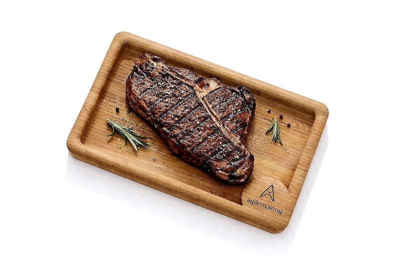 K&ouml;nnte beinhalten: Ein gegrilltes T-Bone-Steak, gew&uuml;rzt mit Pfeffer und Salz, pr&auml;sentiert auf einem rechteckigen Holzservierbrett. Rosmarinzweige und Pfefferk&ouml;rner sind um das Steak angeordnet. Das Brett hat den Text "agilemaking".
