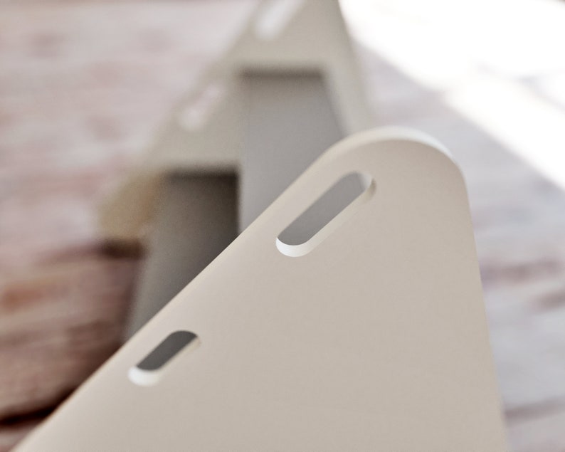 May include: Close-up of a light beige, triangular object with rounded corners and rectangular cut-out handles. The object is in focus, with a second, similar object slightly blurred in the background. The objects appear to be made of a smooth, matte material.