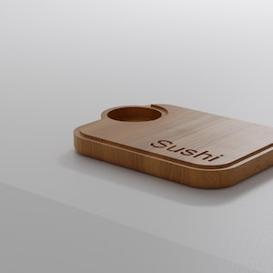 May include: A wooden sushi serving board with a recessed circular area and the word "Sushi" engraved on the surface. The board is rectangular with rounded corners and a raised edge, made of light brown wood.