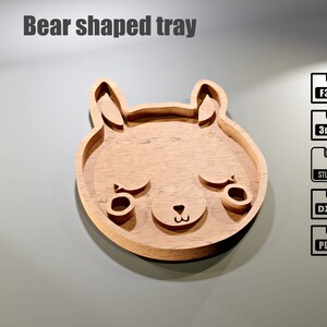 May include: Wooden bear-shaped tray with a light brown finish. The tray features a bear face design with cut-out eyes, nose, and ears. The text "Bear shaped tray" is at the top. Includes file type icons: F3D, 3mf, STL, DXF, and PDF.