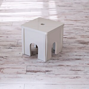 May include: A small, square, white wooden stool with arched cutouts on each side. The stool has a flat top with a single hole in the center. The stool is set on a light-colored, distressed wood floor.