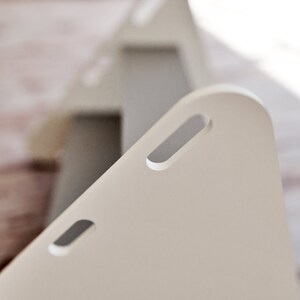May include: Close-up of a light beige, triangular object with rounded corners and rectangular cut-out handles. The object is in focus, with a second, similar object slightly blurred in the background. The objects appear to be made of a smooth, matte material.