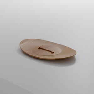 May include: A wooden, oval-shaped tray with a carved, circular indentation and a small, rectangular handle. The tray has a light brown color and a smooth, polished finish. The design is minimalist and modern.