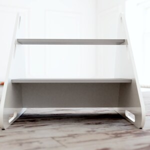 May include: A white two-step stool with gray steps. The stool has a simple design with cut-out handles on the sides. The steps are approximately 12 inches wide, providing a stable platform for reaching higher places.