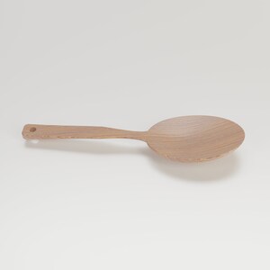 May include: A light-colored wooden spoon with a large, rounded bowl and a long handle. The spoon has a small hole at the end of the handle. The wood grain is visible. This kitchen utensil is designed for stirring and serving.