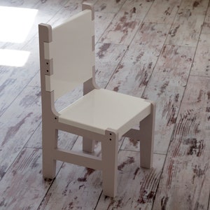 May include: A small, light-colored wooden chair with a white seat and back. The chair has a simple, modern design with a rectangular seat and backrest. The frame is a light gray color, and the chair is set on a wooden floor.