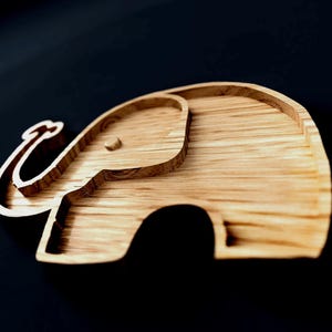 May include: Wooden elephant-shaped tray with a light brown finish. The tray features a detailed outline of an elephant, including its trunk, ears, and body. The tray is made of wood and has a smooth surface. The tray is designed for decorative or functional use.