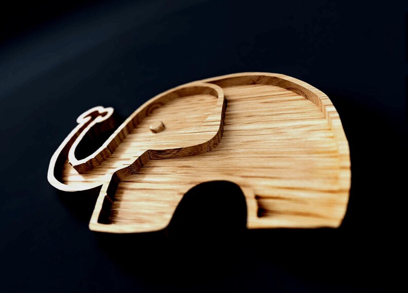 May include: Wooden elephant-shaped tray with a light brown finish. The tray features a detailed outline of an elephant, including its trunk, ears, and body. The tray is made of wood and has a smooth surface. The tray is designed for decorative or functional use.