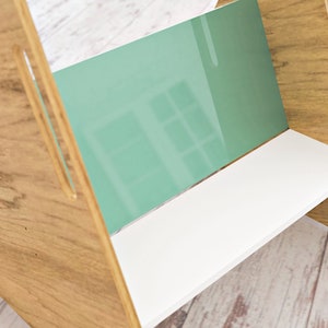 May include: A wooden bookshelf with a light brown finish, featuring a white shelf and a mint green back panel. The design includes cut-out handles on the sides, creating a modern and minimalist aesthetic.