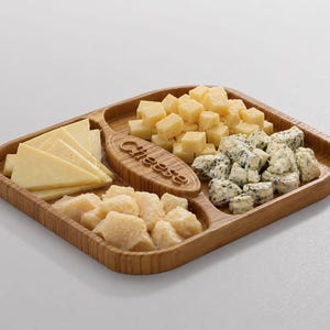 Pode incluir: Uma t&aacute;bua de queijos de madeira com quatro compartimentos, cheia de v&aacute;rios queijos. A t&aacute;bua tem uma pega com a palavra "Cheese" gravada. Os queijos incluem variedades fatiadas, em cubos e esfareladas, em tons de amarelo e branco.