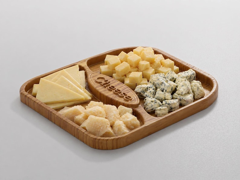 Pode incluir: Uma t&aacute;bua de queijos de madeira com quatro compartimentos, cheia de v&aacute;rios queijos. A t&aacute;bua tem uma pega com a palavra "Cheese" gravada. Os queijos incluem variedades fatiadas, em cubos e esfareladas, em tons de amarelo e branco.