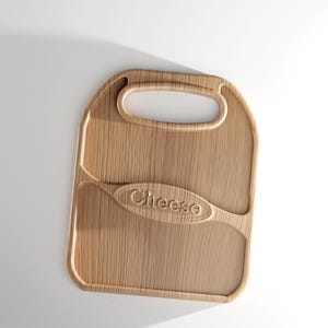 Pode incluir: Uma t&aacute;bua de queijo de madeira clara com uma pega. A palavra "Cheese" est&aacute; gravada na superf&iacute;cie da t&aacute;bua. A t&aacute;bua tem uma forma retangular com bordas arredondadas e um elemento de design curvo.