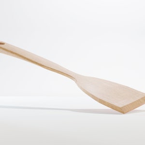 May include: A light-colored wooden spatula with a long handle and a flat, angled head. The spatula has a hole at the end of the handle for hanging. The kitchen utensil is made of a light-colored wood.