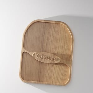 Pode incluir: Uma t&aacute;bua de queijo de madeira castanha clara com cantos arredondados e um design semelhante a uma pega. A palavra "Cheese" est&aacute; gravada na pega. A t&aacute;bua &eacute; feita de madeira e tem um gr&atilde;o natural.