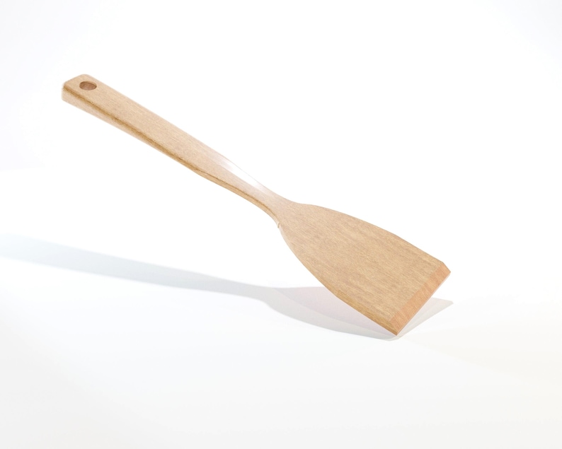 May include: A light-colored wooden spatula with a flat, rectangular head and a long, slender handle. The handle has a small hole at the end for hanging. The spatula is made of a light-colored wood and is designed for cooking and food preparation.