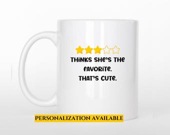 May include: White ceramic mug with a black and yellow design. The text reads "Thinks she's the favorite. That's cute." with four yellow star icons above the text. The bottom of the mug has the text "PERSONALIZATION AVAILABLE" in yellow.